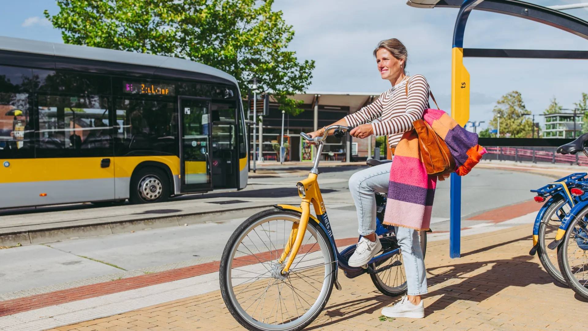 Vrouw op fiets bij bushalte op zonnige dag.