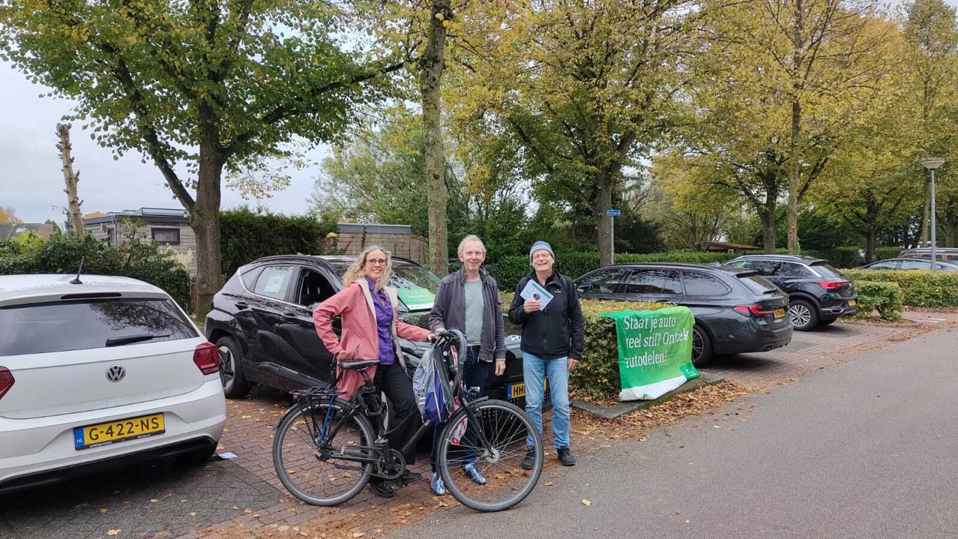 Drie mensen bij een geparkeerde fiets naast auto's, met bomen op de achtergrond en een groen bord. Ze delen folders uit.
