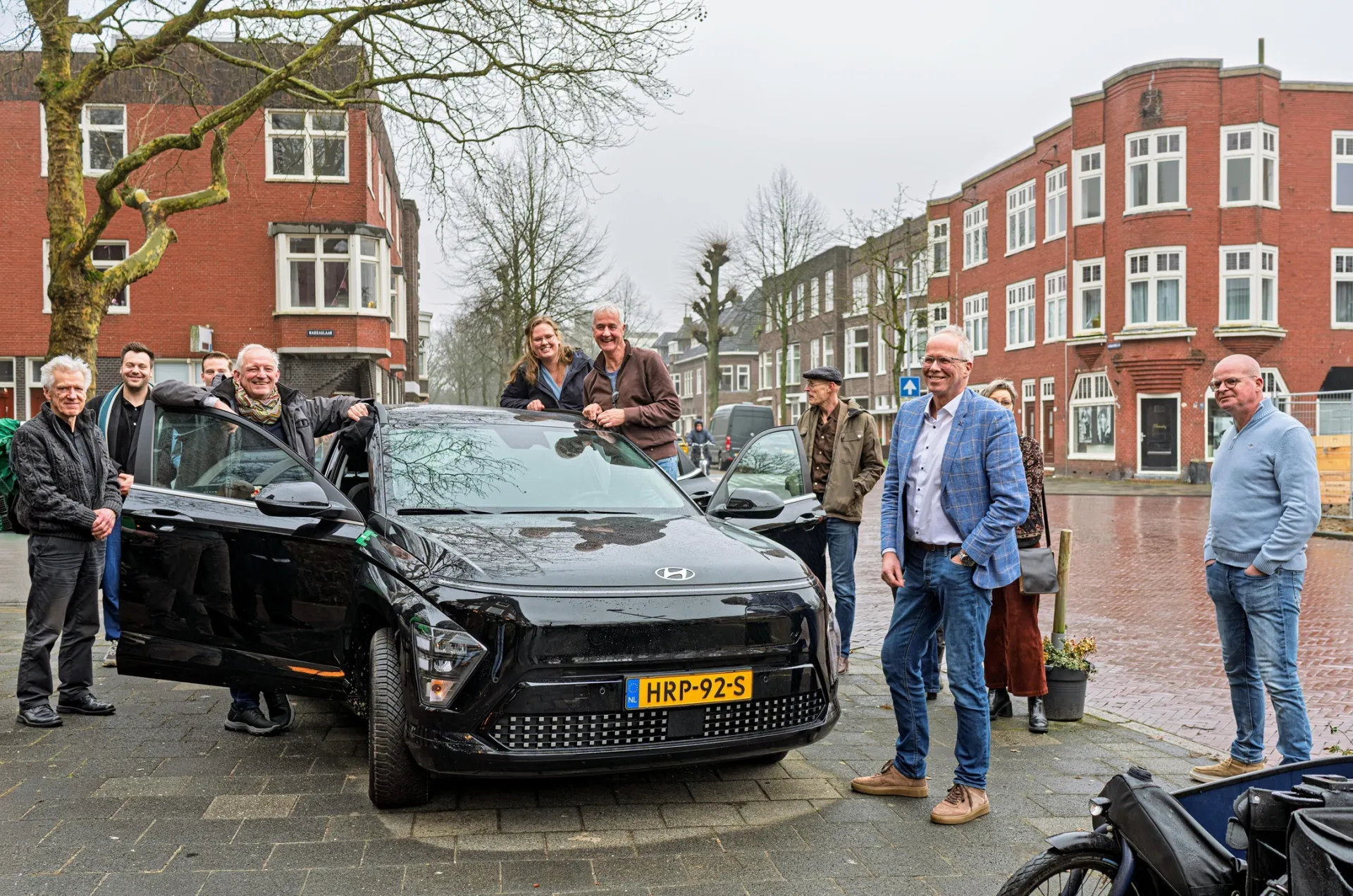 Groep mensen verzameld rond een zwarte auto in een stedelijke straat.