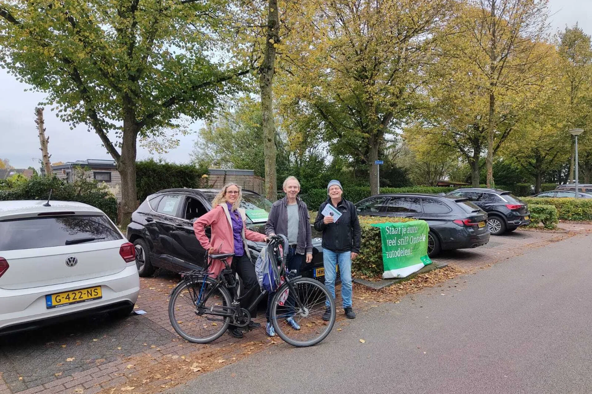 Drie mensen bij een geparkeerde fiets naast auto's, met bomen op de achtergrond en een groen bord. Ze delen folders uit.